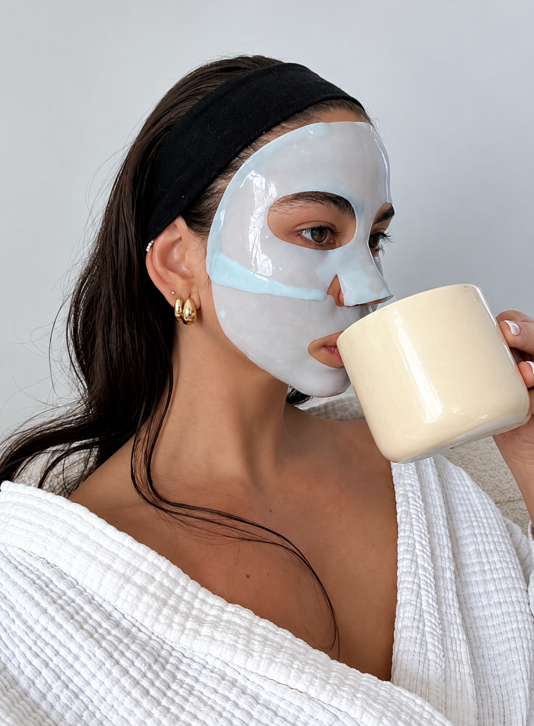 Woman wearing a glossy light blue facial sheet mask and white bathrobe while sipping from a cream-colored mug, with dark hair pulled back by a black headband.
