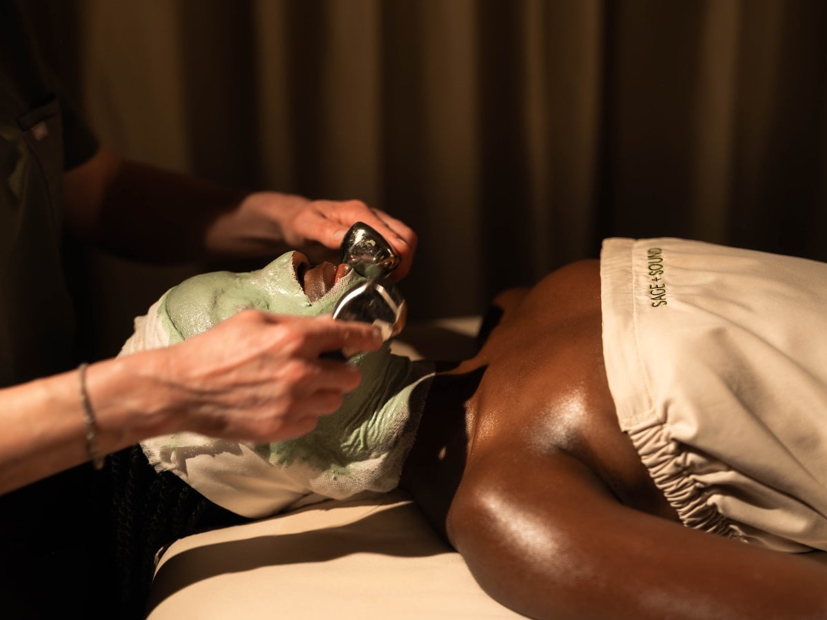 Person receiving a facial treatment with a green face mask while an esthetician uses cooling facial globes to massage the skin.